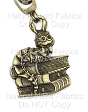 Load image into Gallery viewer, A bronze-colored zipper pull featuring a cartoonish caterpillar wearing glasses and a bow tie, perched atop a stack of books.