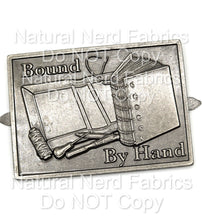 Load image into Gallery viewer, A silver metal plate with the words "Bound by Hand" and an image of a book, tools, and a roll of material. The item is meant for sewing projects as a maker's mark.