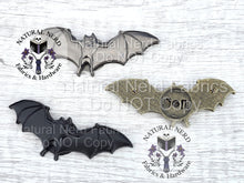 Load image into Gallery viewer, Three metal bat-shaped embellishments in silver, bronze, and black. The bats have detailed wing designs and are arranged on a light wood-grain surface.