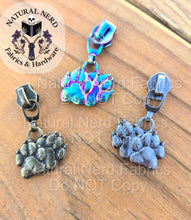 Load image into Gallery viewer, A collection of three detailed zipper pulls designed to look like a pair of canine pay prints. Three finishes are on display: matte rainbow, antique silver, and bronze.