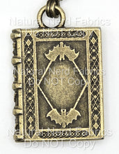 Load image into Gallery viewer, A bronze-toned metal zipper pull shaped like a book with a bat design on the front. The book has a detailed, ornate design with a diamond pattern.
