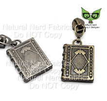 Load image into Gallery viewer, Two metal zipper pulls designed to resemble old, ornate books with a bat motif, one in bronze and one in nickel color.
