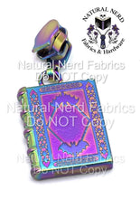 Load image into Gallery viewer, A rainbow-toned metal zipper pull shaped like a book with a bat design on the front. The book has a detailed, ornate design with a diamond pattern.
