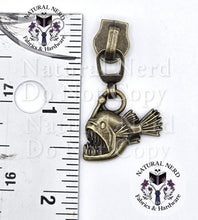 Load image into Gallery viewer, An antique bronze-finished angler fish zipper pull with an open mouth, displayed next to a metal ruler for scale on a white background with brand watermarks.
