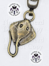 Load image into Gallery viewer, A bronze-toned metal zipper pull shaped like a stingray, featuring detailed eye and mouth carvings, shown against a white background with 'Natural Nerd Fabrics &amp; Hardware' watermarks.
