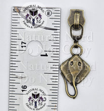 Load image into Gallery viewer, An antique bronze metal common stingray zipper pull shown next to a metal ruler for scale, with a 'Natural Nerd Fabrics &amp; Hardware' watermark overlay.
