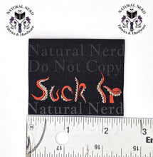 Load image into Gallery viewer, A square black fabric tag with the phrase 'Suck It' embroidered in red and white, shown with a metal ruler for scale and overlaid watermark text from Natural Nerd Fabrics &amp; Hardware.
